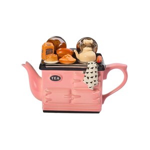 Può includere: Una teiera in ceramica rosa a forma di stufa vintage con un top nero e un'etichetta nera "TEA". La teiera ha un beccuccio e un manico ed è decorata con miniature di cibo, tra cui pane, panini e una piccola teiera.