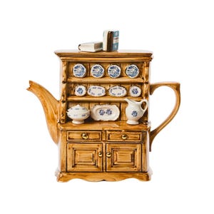 Könnte beinhalten: Eine Keramik-Teekanne in Form einer Holz-Anrichte mit brauner Glasur. Die Teekanne hat einen Ausguss, einen Griff und einen Deckel. Die Anrichte hat Regale mit blauen und weißen Tellern und eine Schublade mit goldenen Griffen. Auf der Anrichte liegen zwei Bücher.