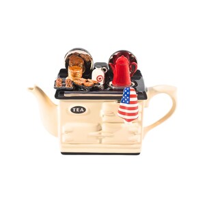 May include: A cream-coloured teapot shaped like a vintage stove with a red, white and blue American flag hanging on the side. The stovetop has a red coffee pot, a small milk jug, and a plate of pancakes.