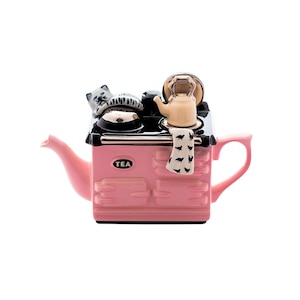 Puede incluir: Una tetera rosa con forma de estufa vintage con una placa de cocción negra, un paño de cocina blanco y negro y una figurita de gato en la parte superior. La tetera tiene un asa y un pico negros y dice "TEA" en la parte delantera.