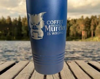 May include: A blue insulated tumbler with a silver lid, featuring an etched image of a grumpy cat holding a coffee cup. The text on the tumbler reads "Coffee Because Murder Is Wrong."