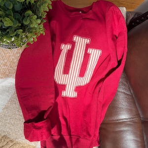 May include: A crimson sweatshirt with a large white and red striped "IU" logo. The long-sleeved crewneck top is folded, showing the ribbed cuffs. The sweatshirt is made of a soft material, suitable for casual wear.