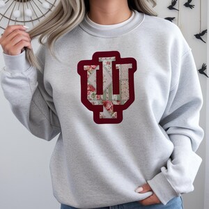 May include: A light grey sweatshirt with a maroon and floral "IU" logo. The logo is a stylized monogram with a floral pattern and a maroon outline. The sweatshirt has long sleeves and a crew neck.
