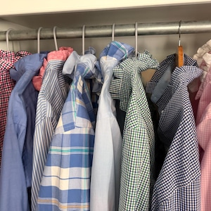 May include: A collection of button-down shirts hanging on a metal rod, featuring various patterns including red and blue gingham, solid light blue, vertical blue stripes, blue plaid, solid white, green gingham, blue check, and light pink gingham.