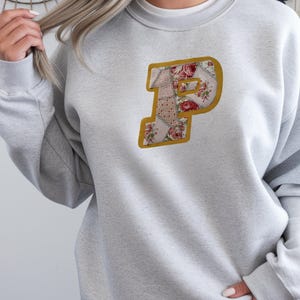 May include: A light gray sweatshirt featuring a floral letter "P" applique. The letter is outlined in gold and filled with a patchwork design of floral patterns in red, pink, and white. The sweatshirt has long sleeves and a crew neck.