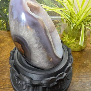 May include: A polished, egg-shaped agate geode featuring a smooth, translucent lavender and grey exterior with a hollow center lined with sparkling, light-colored crystals, resting securely on a matte black, circular lotus-shaped stand.
