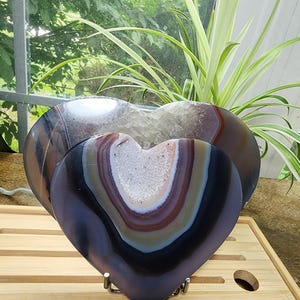 May include: Two heart-shaped agate stones with polished dark brown and grey banded surfaces. The center of each stone features a hollow geode cavity filled with small, sparkling white crystals. The stones are displayed on a metal stand.