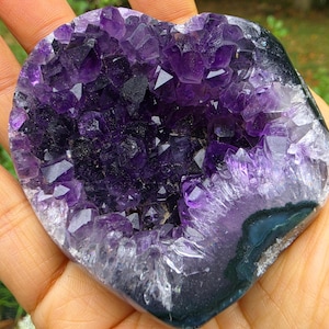 May include: A heart-shaped amethyst geode featuring deep purple, faceted crystal points clustered within a dark, polished stone border. The natural mineral specimen displays a vibrant violet hue and a jagged, crystalline texture throughout the center.