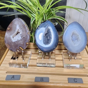 May include: Three teardrop-shaped agate geodes displayed on clear acrylic stands. The left geode is brown with a white center, the middle is dark blue with a deep cavity, and the right is light blue with a crystalline core. Labels A, B, and C are present.