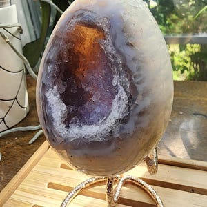 May include: An egg-shaped geode featuring a smooth, pale, translucent outer shell and a deep, hollow center filled with sparkling, dark purple amethyst crystals and warm, amber-colored mineral deposits, supported by a gold-toned metal stand.