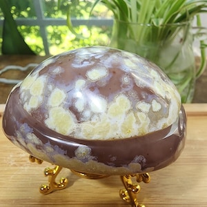 May include: A polished, dome-shaped flower agate stone featuring a smooth, mauve-brown base with creamy, pale yellow floral-like inclusions. The stone is displayed resting on a small, ornate, gold-colored metal tripod stand with curved legs.