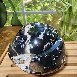 May include: A polished black stone bowl with a matching lid, featuring natural white and tan mineral inclusions and crystalline formations. The surface is smooth and glossy, showcasing organic patterns of white speckles and jagged veins.