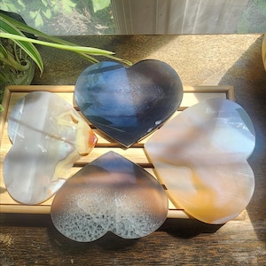May include: Four heart-shaped polished stone coasters with smooth surfaces. One is dark charcoal, one is translucent white with orange veins, one is pale cream with yellow swirls, and one is a smoky grey with a textured crystalline center pattern.