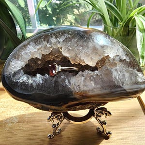 May include: A silver ring featuring a deep red oval gemstone flanked by two small clear stones, resting inside a large, hollowed-out geode with dark outer layers and a sparkling, crystalline interior, supported by a decorative metal stand.