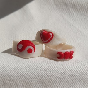 Könnte beinhalten: Drei weiße Ringe mit roten Designs. Der erste Ring hat ein rotes Yin-Yang-Symbol, der zweite Ring hat ein rotes Herz und der dritte Ring hat ein rotes Dreifachmond-Symbol.