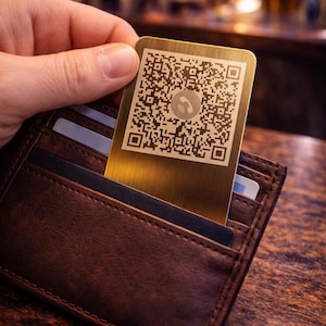 May include: A gold-colored metal business card with a QR code and phone icon is held above a brown leather wallet. The card is rectangular and features a detailed black and white QR code.