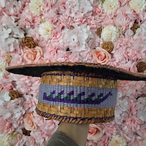 May include: A woven hat with a wide brim, featuring a band of purple and green geometric design. The hat is made of natural-coloured woven material and has a dark rim. The background is a wall of pink and white flowers.