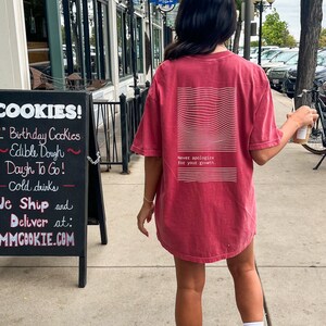 Puede incluir: Una mujer con una camiseta roja con texto blanco que dice "Nunca te disculpes por tu crecimiento" caminando por una acera. La camiseta tiene un dise&ntilde;o gr&aacute;fico blanco en la espalda. Lleva una taza de caf&eacute; en la mano derecha.