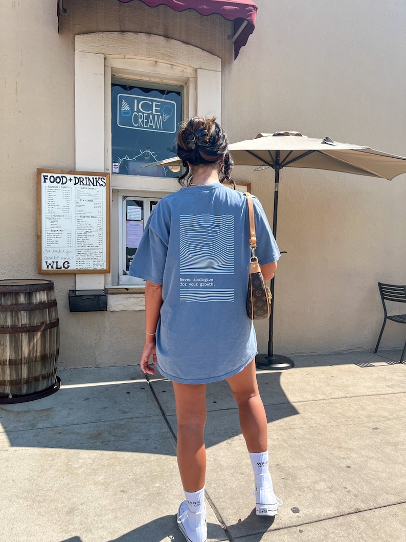 Puede incluir: Una mujer lleva una camiseta azul claro con el texto "Nunca te disculpes por tu crecimiento" en la espalda. Lleva calcetines blancos y zapatillas blancas. Est&aacute; de pie frente a un edificio con un cartel que dice "Helado".