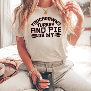 May include: White t-shirt with brown text that reads "Touchdowns Turkey and Pie Oh My".