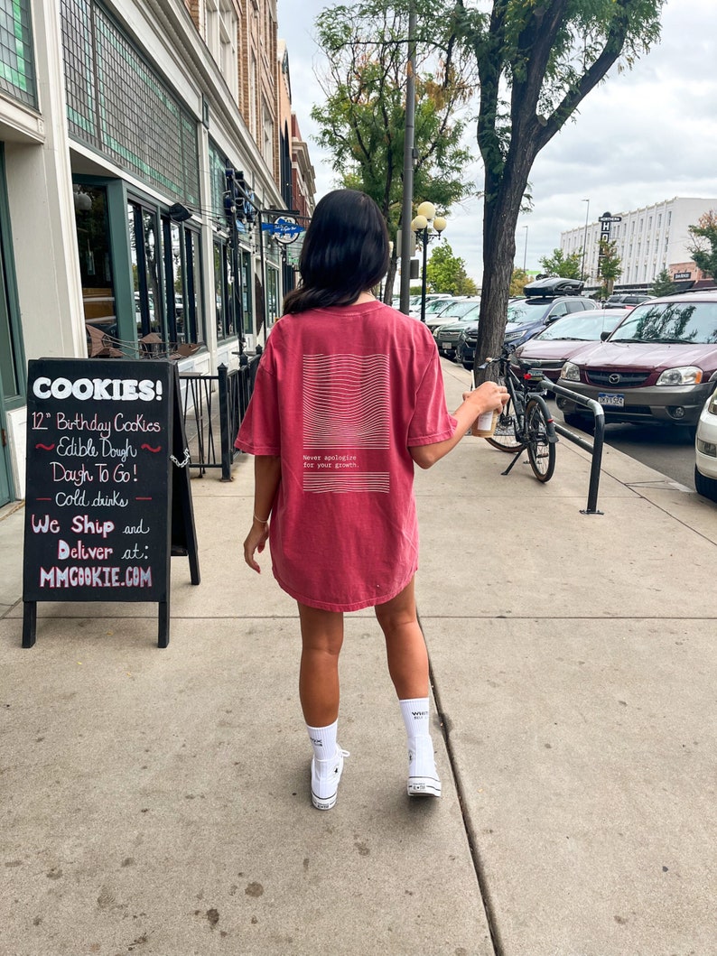 Puede incluir: Una mujer con una camiseta roja con texto blanco que dice "Nunca te disculpes por tu crecimiento" caminando por una acera. La camiseta tiene un dise&ntilde;o gr&aacute;fico blanco en la espalda. Lleva una taza de caf&eacute; en la mano derecha.