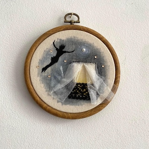 May include: Embroidered artwork in a wooden hoop. The design features a black silhouette of a figure flying, a tent, and a night sky with a pearl and sparkling accents. The hoop has a metal clasp.