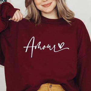May include: A maroon sweatshirt with the word "Amour" written in white cursive with a small heart symbol next to it.