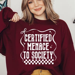 May include: A maroon sweatshirt with the text "CERTIFIED MENACE TO SOCIETY" in white, with a decorative border and a checkered pattern at the bottom. The sweatshirt is long-sleeved and has a crew neck.