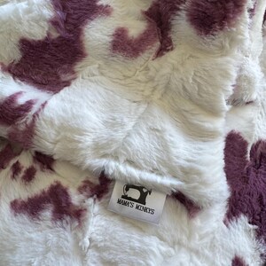 May include: A soft, white blanket with irregular, deep purple patches. The blanket has a fluffy texture. A label with the text "MAMA'S MINKYS" and a sewing machine logo is visible.