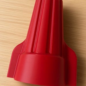 Pode incluir: Um conector de fios de plástico vermelho brilhante com um corpo cilíndrico cônico e canelado, apresentando duas abas laterais largas e planas para torção manual. O componente possui um acabamento liso e uma base oca para inserção.