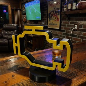 Pode incluir: Um item decorativo em forma de motor, preto e amarelo. O item está em uma base preta e fica em um bar de madeira. O fundo inclui uma TV, uma placa de néon que diz "Bereex" e um pôster vintage.