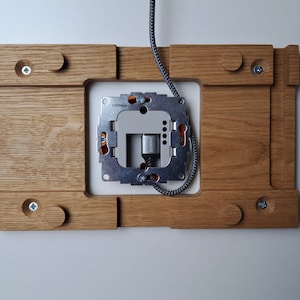 May include: A wooden wall plate with a metal electrical outlet box mounted in the center. The plate has a light brown finish and is designed to be installed on a wall.
