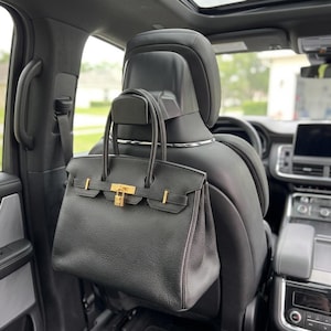 Op de afbeelding: Een zwarte leren handtas met goudkleurige hardware, inclusief een klein hangslot en gespriemen, hangend aan een zwarte plastic haak bevestigd aan de hoofdsteun van een autostoel. De handtas heeft een getextureerd oppervlak en twee handvatten.