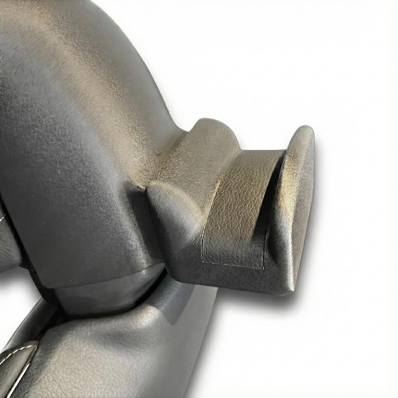 K&ouml;nnte beinhalten: Nahaufnahme eines Teils eines dunkelgrauen Fahrzeuginnenraums. Das Teil hat eine strukturierte Oberfl&auml;che und ein gebogenes Design mit einem markanten, abgewinkelten Vorsprung. Das Material scheint ein haltbarer Kunststoff oder ein synthetisches Material zu sein.