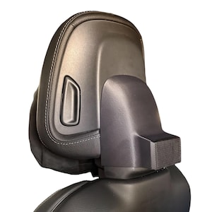 May include: A black car seat headrest with a curved design. The headrest is made of leather with white stitching and a plastic component. The headrest has a rectangular indentation and a textured section.