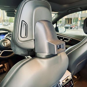 以下が含まれることがあります： 黒い車のシートヘッドレストのクローズアップ。内蔵された調整可能なコンポーネントがあります。ヘッドレストは革製で、洗練されたモダンなデザインです。背景には、ステアリングホイールとダッシュボードを含む車のインテリアが見えます。