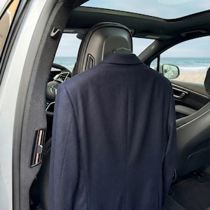 May include: A navy blue blazer hanging in a car, with a beach visible through the open door. The blazer is a classic style, with a notched lapel and two buttons. The car's interior is black leather.