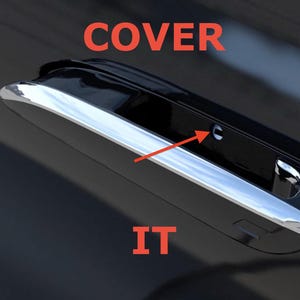 May include: A close-up of a black car door handle with a chrome accent. The word "COVER" is written in red above the handle, and the word "IT" is written in red below the handle. An arrow points to a small hole.