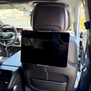 May include: A black tablet is attached to the back of a car seat. It's held by a silver metal bracket. The car's interior is visible, including the steering wheel and dashboard.