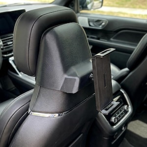 May include: A black plastic tablet mount attached to the back of a car headrest. The mount features an adjustable rectangular frame with a central support arm, designed to hold a mobile device securely in place for rear-seat viewing.