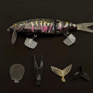 May include: A multi-jointed fishing lure with a black, yellow, and pink scale pattern, featuring a clear plastic diving lip and two treble hooks with white protective covers. Below are four interchangeable components: a clear lip, two tail fins, and a black fin.