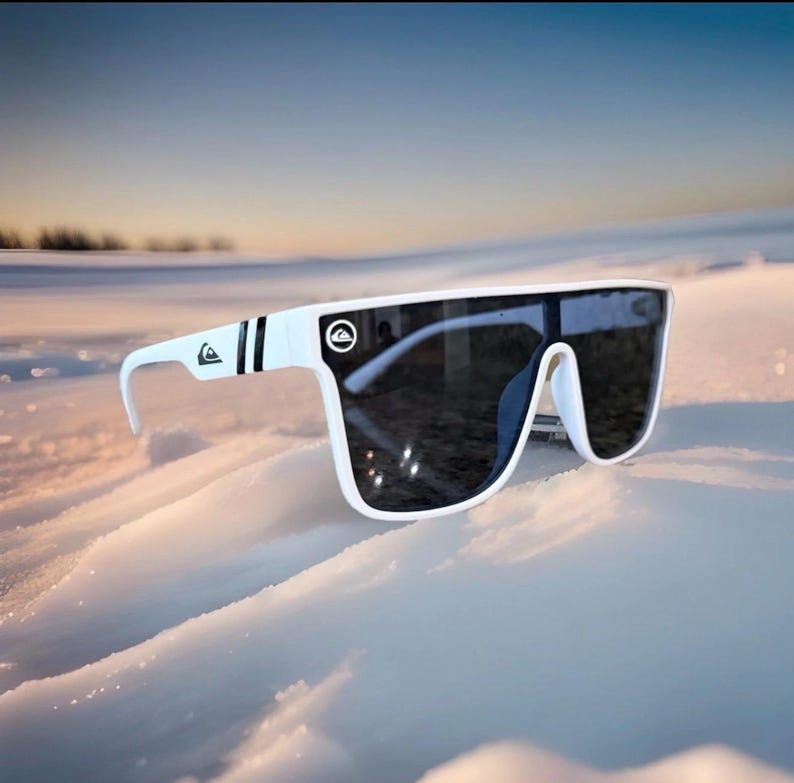 May include: White sunglasses with dark lenses. The sunglasses feature a thick white frame with black stripes on the arms and a logo. They are set against a snowy backdrop.