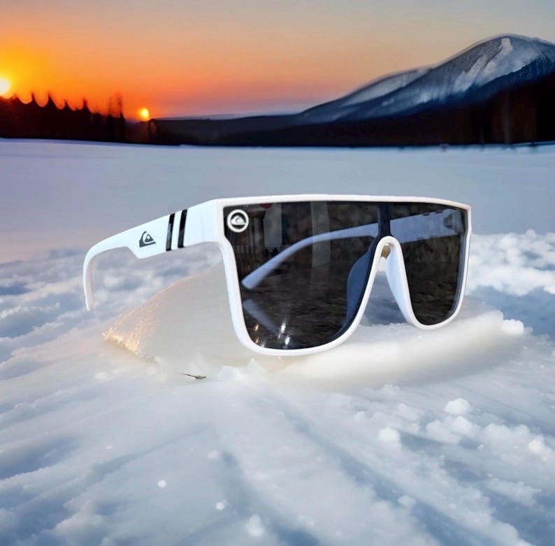 May include: White sunglasses with a wide frame and dark lenses. The sunglasses have a logo on the temple and a black stripe. The background shows a snowy landscape with a sunset.
