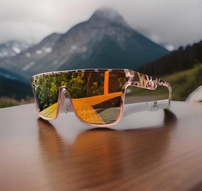 K&ouml;nnte beinhalten: Sonnenbrille im Shield-Stil mit einem hellrosa und braun gemusterten Leoparden-Rahmen. Das gro&szlig;e Ein-Scheiben-Design verf&uuml;gt &uuml;ber eine reflektierende, goldfarbene Verspiegelung, die die Umgebung klar widerspiegelt. Das Quiksilver-Logo ist sichtbar.