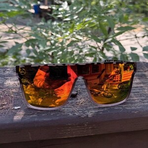 K&ouml;nnte beinhalten: Sonnenbrille mit flachem Rahmen im Schildpattmuster und reflektierenden, orange get&ouml;nten Gl&auml;sern. Die Gl&auml;ser haben eine spiegelnde Oberfl&auml;che, die die Umgebung reflektiert, und ein kleines Logo ist auf dem linken Glas zu sehen.