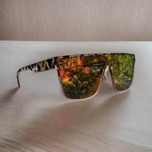 K&ouml;nnte beinhalten: Sonnenbrille im Shield-Design mit einem durchgehenden Glas in reflektierender Gold- und Gr&uuml;noptik. Der Rahmen ist an den B&uuml;geln mit einem schwarz-wei&szlig;en Tiermuster versehen, das zudem ein kleines wei&szlig;es Quiksilver-Logo aufweist.