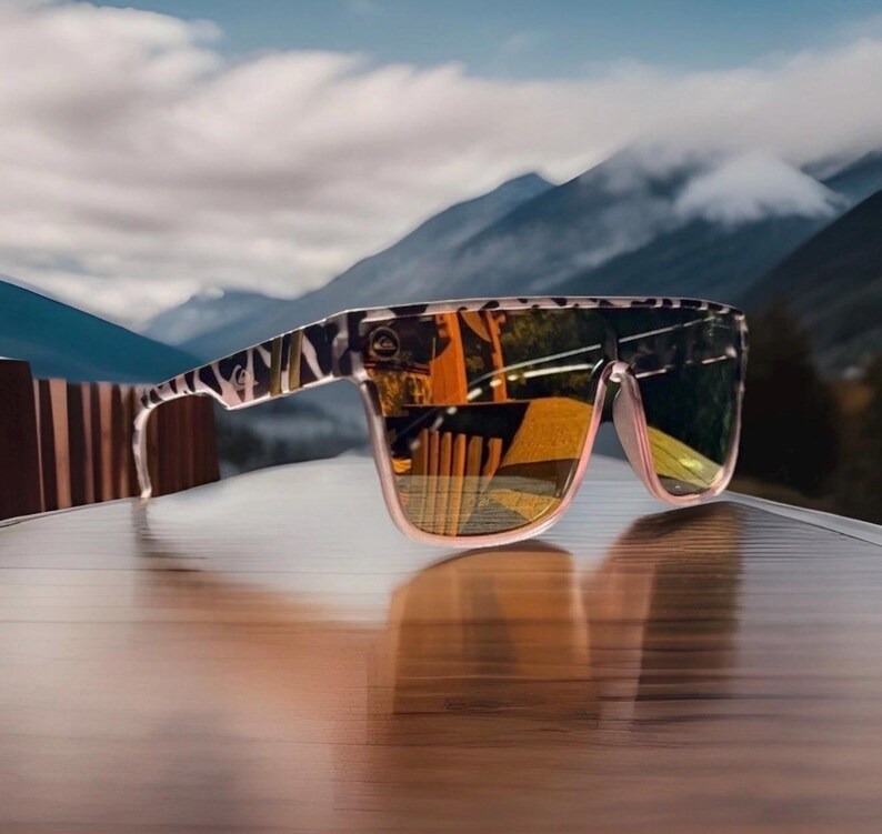 K&ouml;nnte beinhalten: Sonnenbrille mit flachem Rahmenoberteil und einem schwarz-wei&szlig;en Tiermuster auf den B&uuml;geln und der Vorderseite. Die Gl&auml;ser sind reflektierend mit einem warmen Orange- und Gelbstich und zeigen eine Spiegelung einer Holzstruktur.
