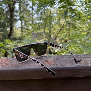 K&ouml;nnte beinhalten: Sonnenbrille mit flachem Rahmen und einem schwarz-beigen Leopardenmuster an den B&uuml;geln und den vorderen R&auml;ndern. Die Gl&auml;ser sind dunkel get&ouml;nt und gehen am unteren Rand des Rahmens in einen helleren, durchscheinenden Rosaton &uuml;ber.