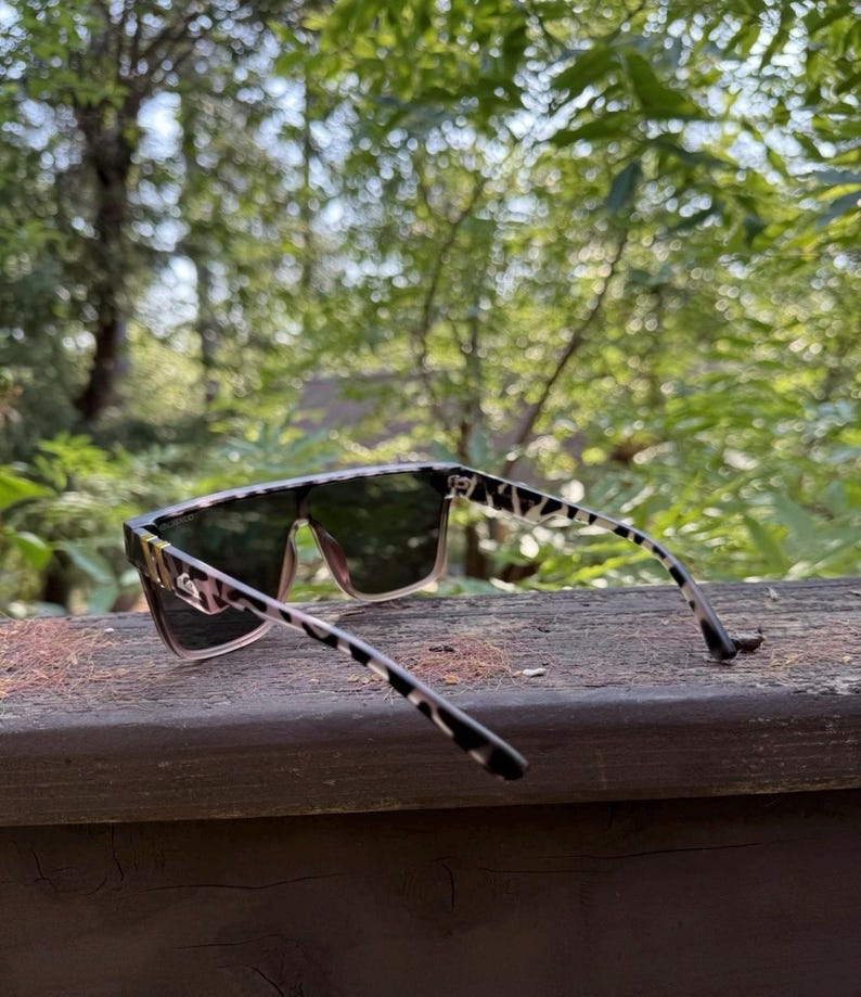 K&ouml;nnte beinhalten: Sonnenbrille mit flachem Rahmen und einem schwarz-beigen Leopardenmuster an den B&uuml;geln und den vorderen R&auml;ndern. Die Gl&auml;ser sind dunkel get&ouml;nt und gehen am unteren Rand des Rahmens in einen helleren, durchscheinenden Rosaton &uuml;ber.