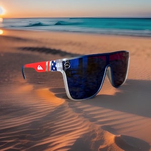 May include: Large, wraparound sunglasses with a patriotic design. The frames are clear with red and blue accents, and the arms feature a red and white pattern with blue stars. The lenses are dark, and the sunglasses are resting on a sandy beach.